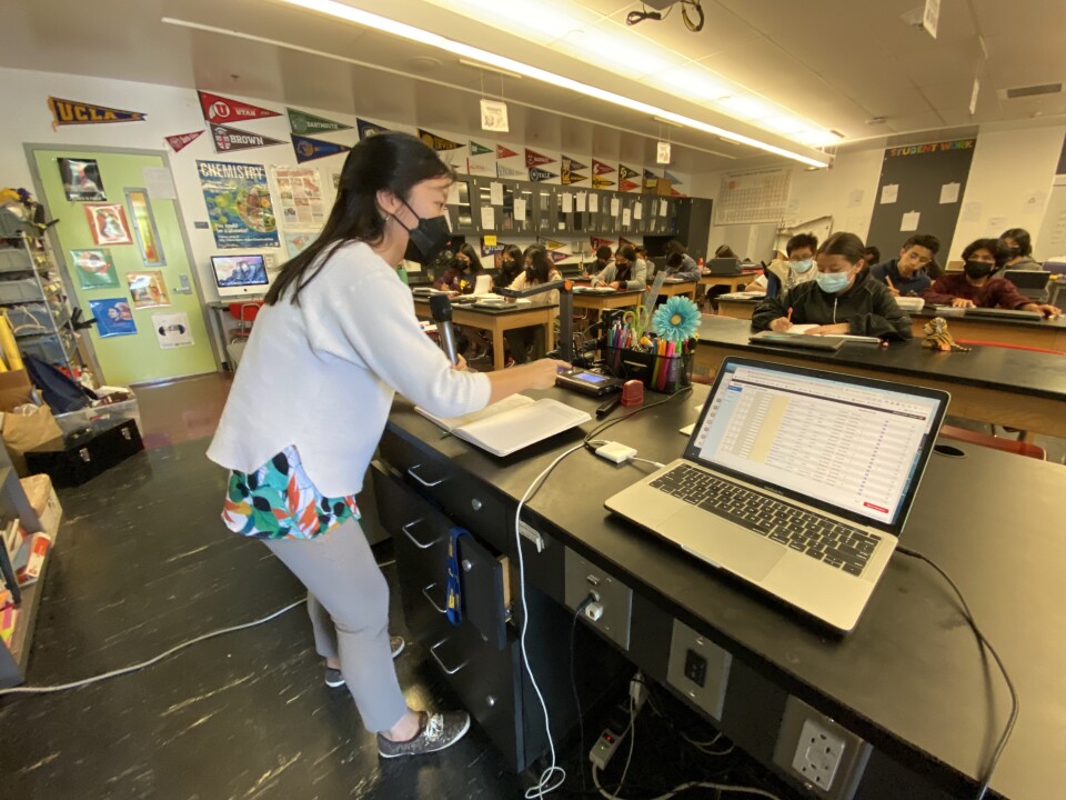 A woman in a white sweater stands at the front of a classroom with her laptop open. She holds a black microphone as she leads a class with students looking on in the background, seated at high-top chemistry benches.