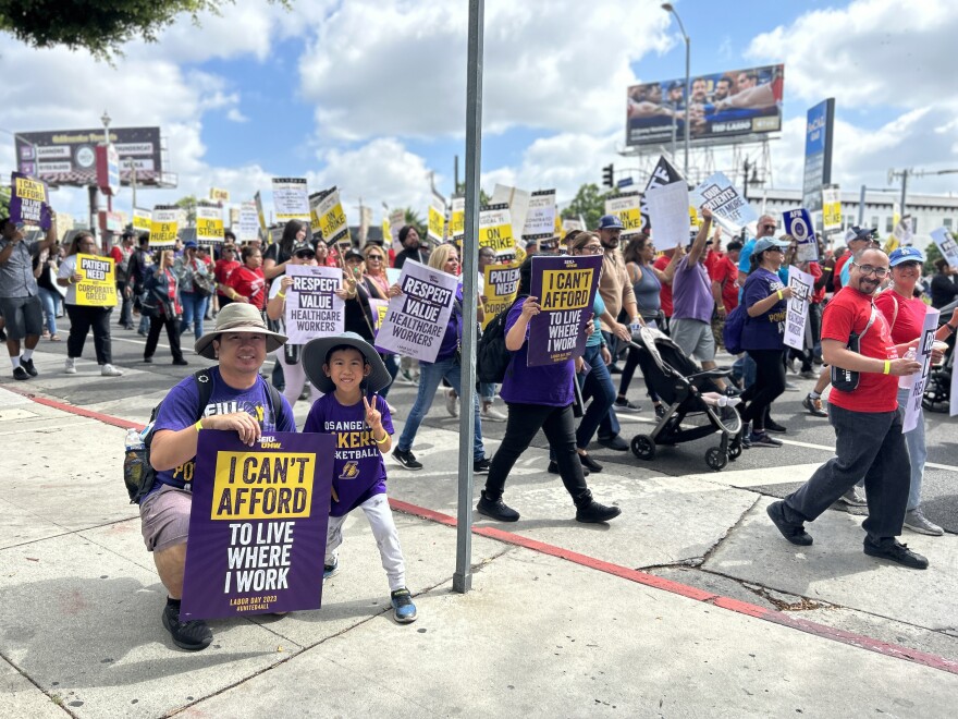 A light-skinned man wearing a tan wide-brimmed hat crouches with on a sidewalk with a sign that reads "I can't afford to live where I work." His young son also wears a wide-brimmed hat and stands to his father's left holding his hands up in a peace sign. They both wear purple shirts. Protestors march in the background.