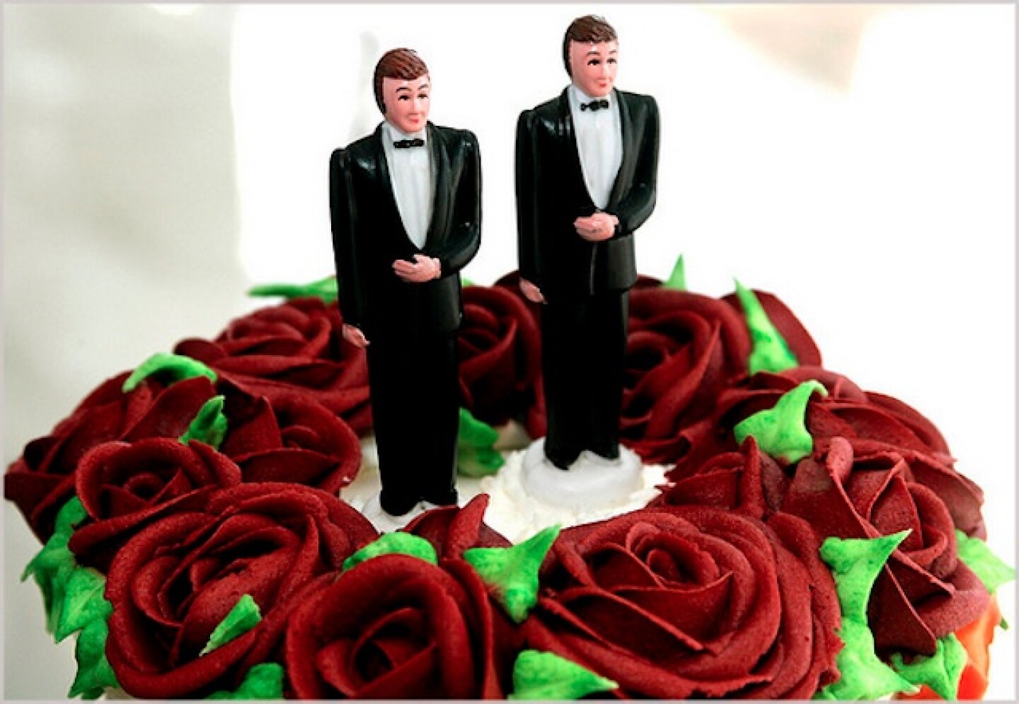 Two plastic grooms are cake toppers on a white wedding cake with red roses.