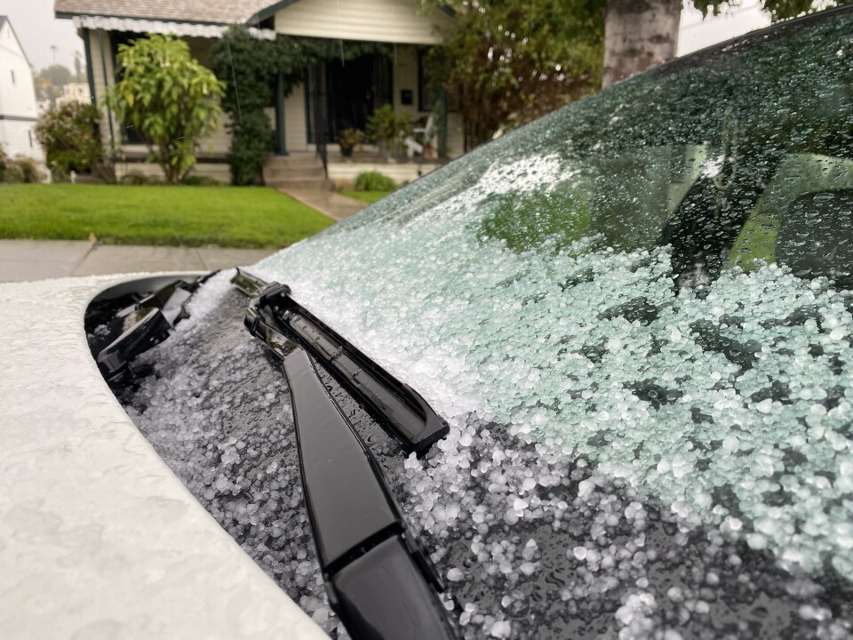 Hail covers the hood of a car parked on a street in front of a bungalow