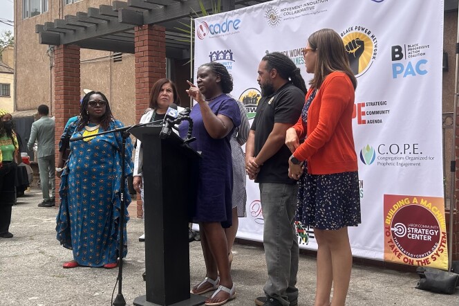 Six people, five women and one man, are lined up behind a black podium with microphones attached. The woman in the center is speaking directly into the microphone with her left hand raised, her finger pointing out towards the unseen audience. They're standing in front of a white banner with several different organization's logos on it.