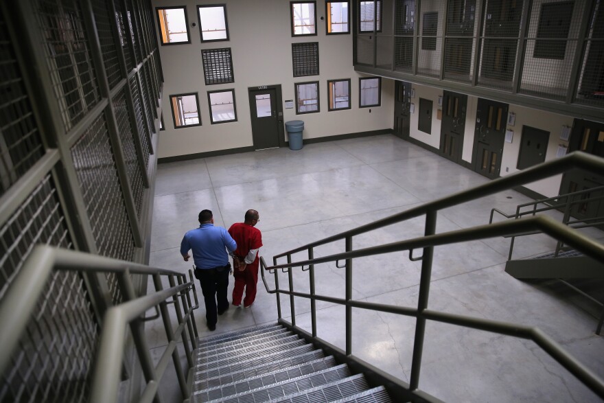 An image of a detainee being escorted by a guard down a flight of stairs inside an immigration detention center. Cells flank the staircase on both sides.