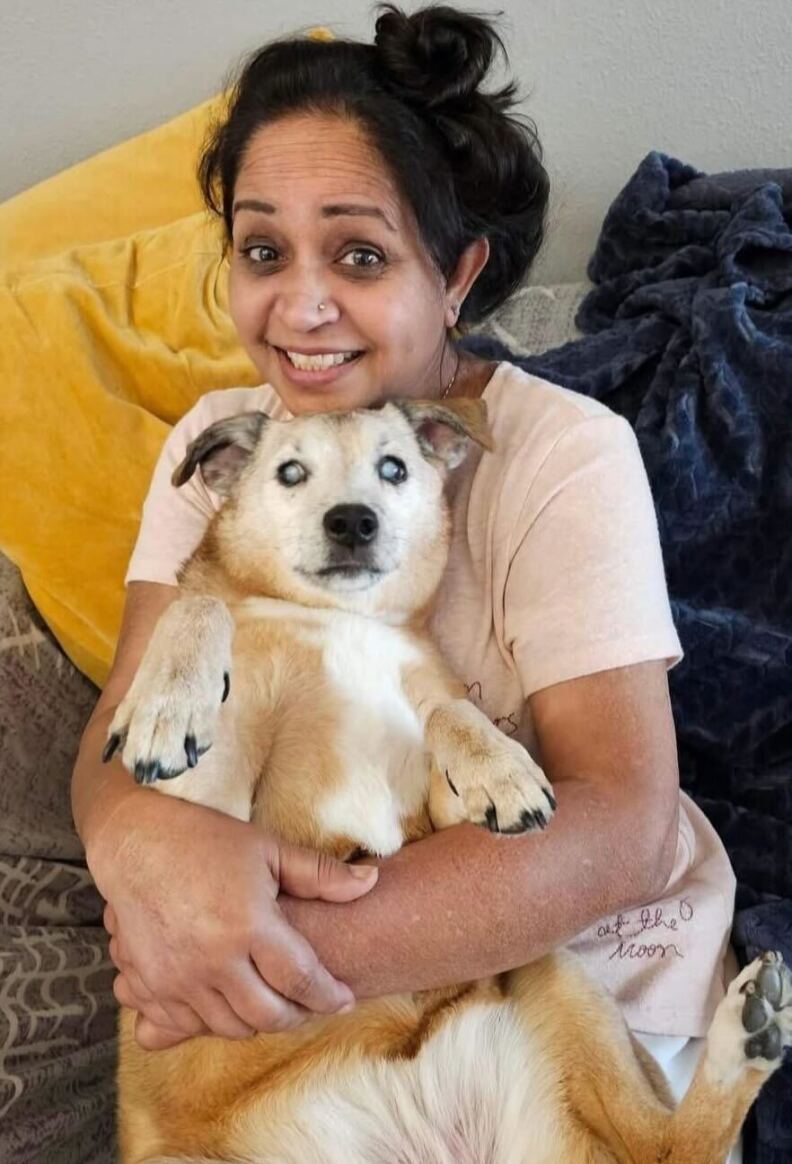 A South Asian woman with dark hair and wearing a pink t-shirt cuddles a dog.