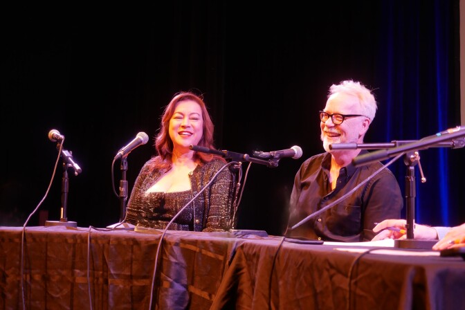 Jennifer Tilly, a white woman and Adam Savage, a white man with short gray hair and glasses, sitting at the judges table and laughing