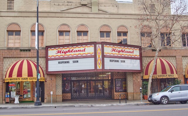 A view of Highland Theatre from across the street. A marquee reads "REOPENING SOON" in the space for changeable lettering, and a permanent sign above that reads "Highland" in red script, with a zig zag border in red and yellow. Red and yellow striped awnings on either side of the sign read "Magda's Hair Cuts" on the left and "Lovely Nails" on the right. Metal gates in front of the front doors are closed.