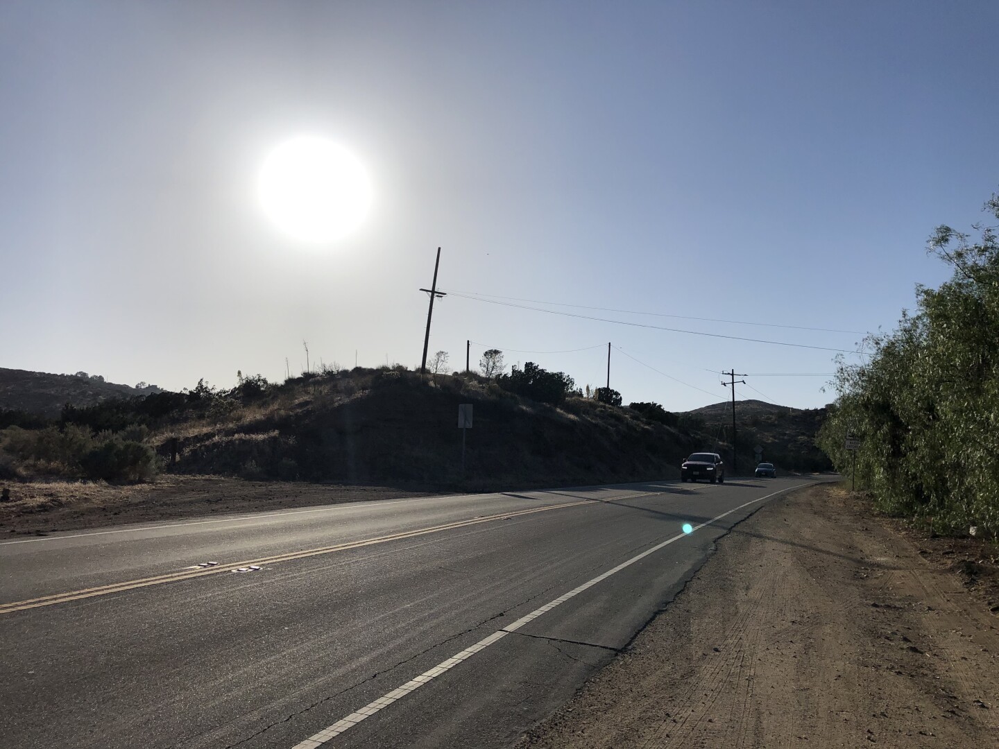 An empty street is pictured with a bright and hot sun bearing down overhead. Three power poles with wires are beside the road.