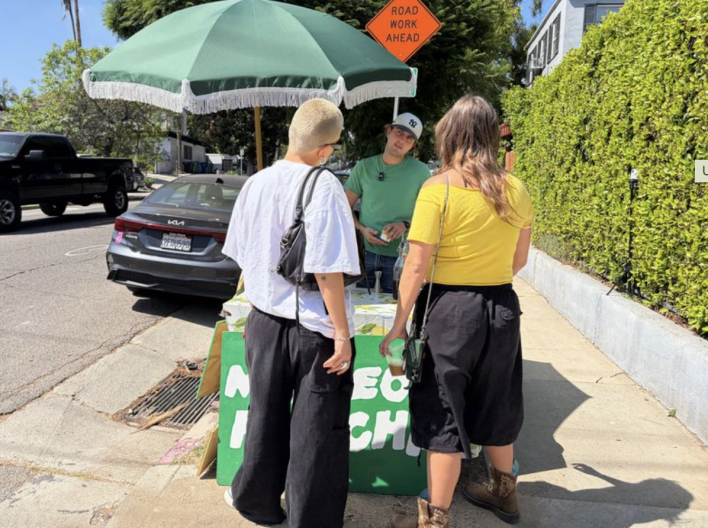 A light skinned man and woman gather at a sidewalk stand with a green umbrella, buying drinks from a vendor on a sunny street.