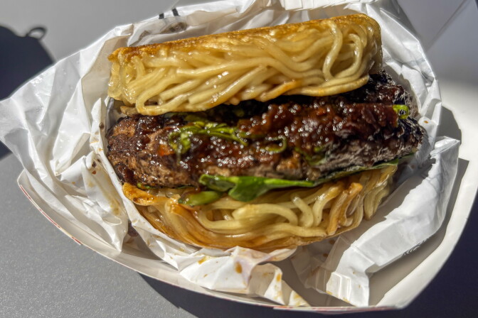A close-up of a ramen burger — a beef patty glazed in dark sauce with arugula, sandwiched between two seared noodle buns — resting in white food paper.