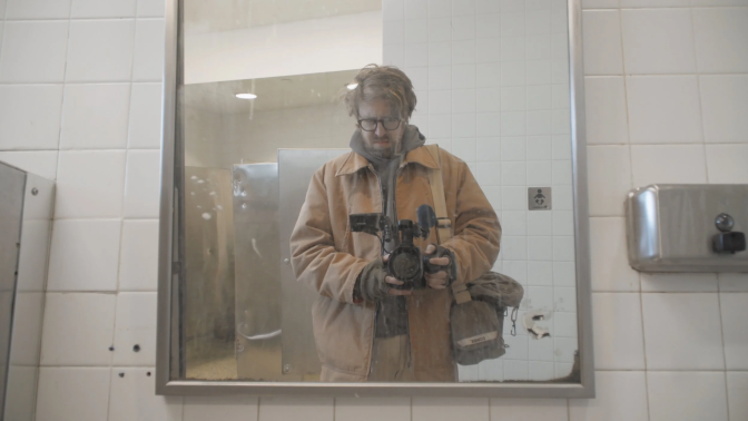 A male presenting man with glasses in a tan jacket looks at himself in a bathroom mirror with a camera pointed towards the mirror.
