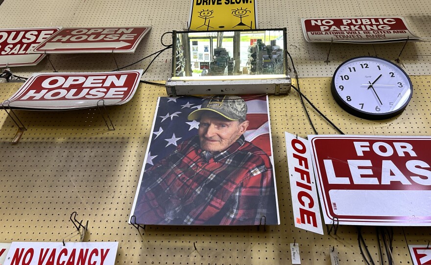 A portrait of an older man, wearing a red plaid flannel button-up a camouflage U.S. Army baseball hat, is displayed towards the ceiling of a wall. The photo is surrounded by different red and white signs, including those that read "open house" "for lease" and "no vacancy." An analog clock is on the right, with the time reading a little before 1:30.