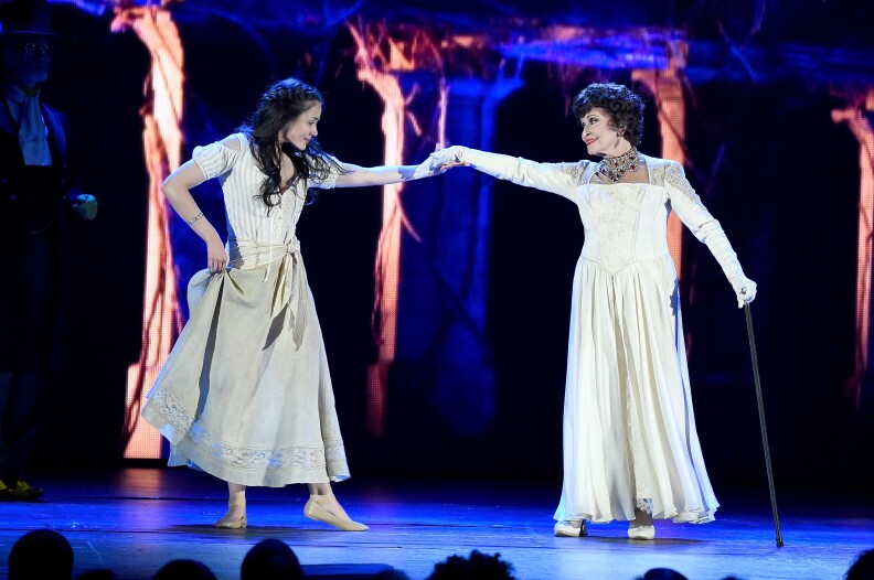 Two women — a younger one on the left, an older one on the right — are locking hands as they perform on stage.