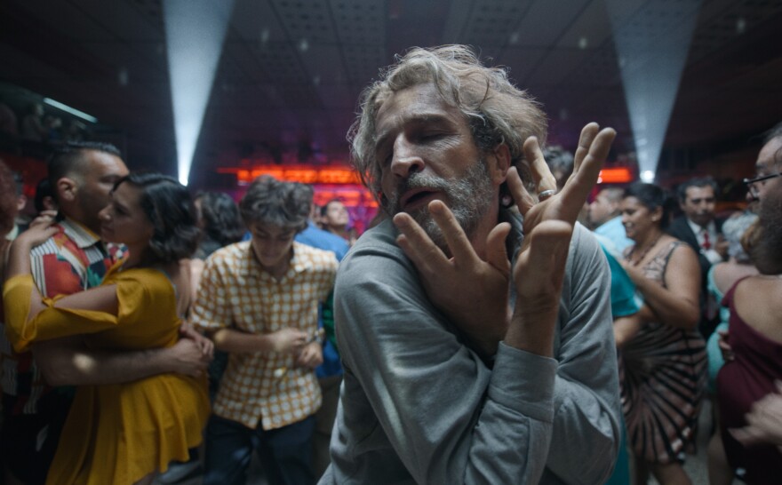 Daniel Gimenez Cacho stands on the dance floor in a crowded club. He wears a light blue button-up shirt with long sleeves. His arms are crossed over the front of his body at the wrists as he bends slightly forward. His hands reach up towards his face, fingers spread out. Cacho's eyes are closed and his mouth is opened slightly. He seems to be dancing. His hair is gray and shaggy, falling loosely below his ears. Cahco has a gray beard that is short but untrimmed. The crowd surrounding him are men and women of different ages, wearing colorful clothing all dancing, some with partners and some by themselves. The back wall is lit up with orange and purple lights with two white beams of light flanking each side shining towards the ceiling.