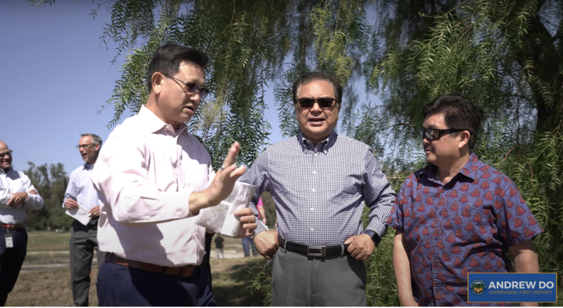 Three Asian men stand talking in a what appears to be a park park, all are wearing sunglasses