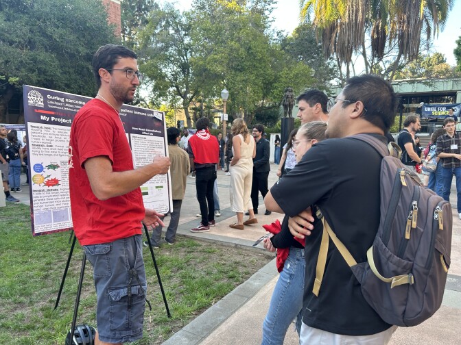 A man with light skin tone stands before a scientific research poster in a courtyard. In front of him, three other people listen as he speaks. In the background, people pause to look at other posters. 