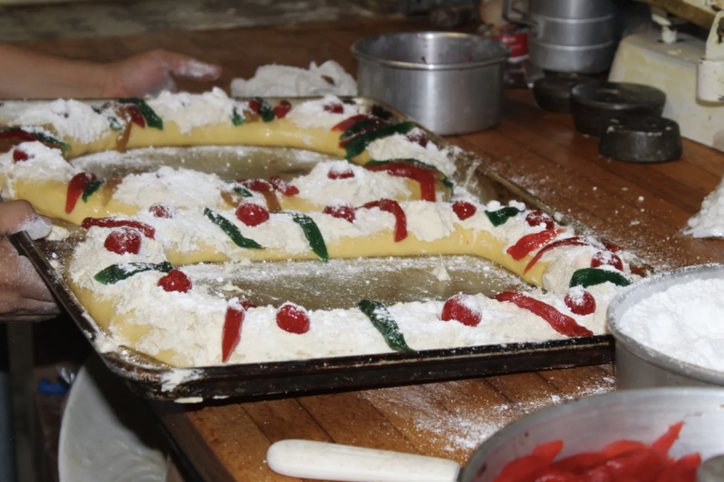 A close up of dough in the shape of an oval ring, decorated with white icing, red cherries and other red and green sweet additions. There are two rings on trays next to each other on a wooden counter, with icing sugar sprinkled on the counter as if the rings had just been dusted