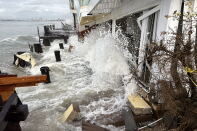 Waves surge against a white waterfront building, splashing up its wall and surrounding broken wooden decking and posts.