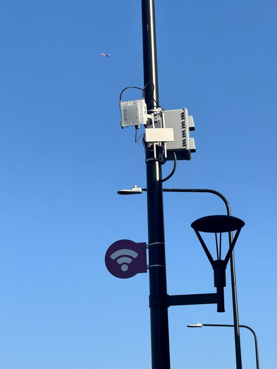 A black street light pole has white boxes attached to it. Off to the left is a small, blue sign with a wi-fi symbol on it. 