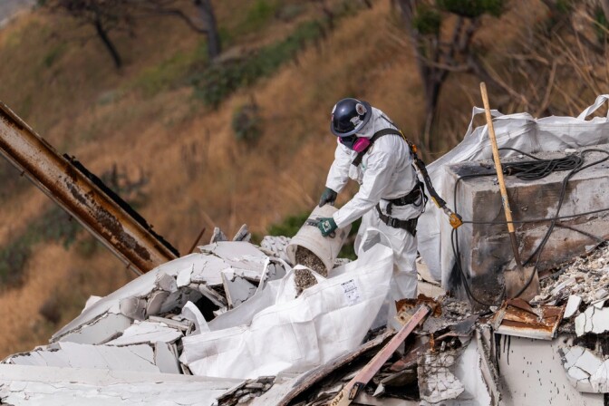 A man wearing a white safety suit clears debris by hand from a hillside property that burned in a fire. 