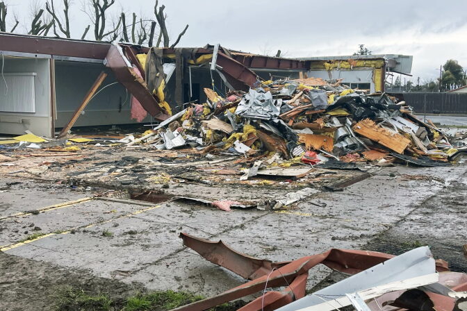 A single-story school building with its roof torn open and walls partly collapsed, leaving a large pile of splintered lumber, twisted metal, and pink and yellow insulation on the wet pavement. Bent red steel beams lie in the foreground, and nearby trees are stripped of their branches.