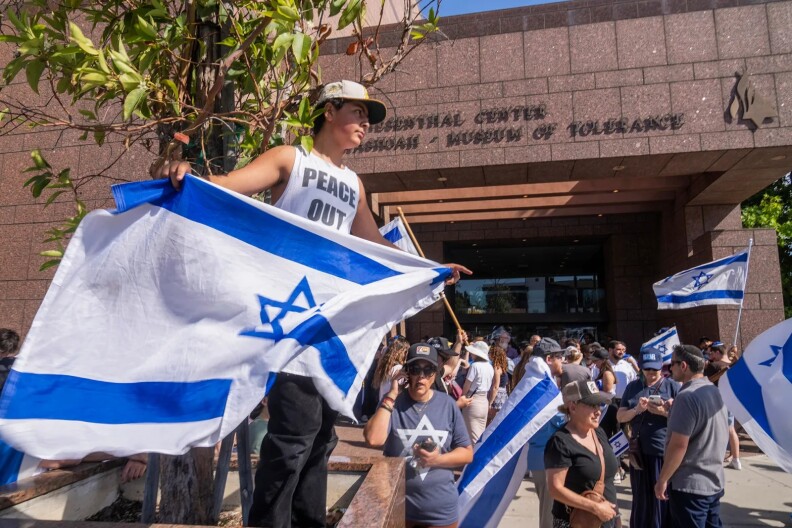 A young male-presenting person stands in front of crowd holding an Israeli flag at a protest at the Museum of Tolerance