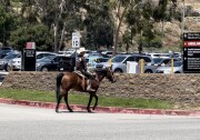 A man with a sombrero and dark clothing rides a horse near a parking lot on a sunny day.