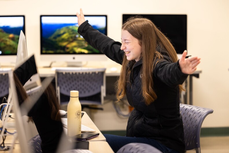 A woman in a black sweater with long auburn hear stretches out her arms as she smiles with her eyes closed. She's in a room full of desks and computer monitors.