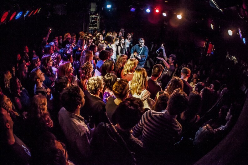 Two men stand on a stage as an audience surrounds them learning how to sing songs.