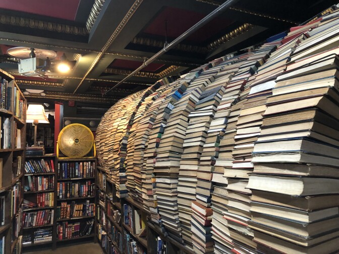 Rows of books sitting on top of a short bookshelf all arranged with their spines facing and created a semi-circle or curved effect on the right side of the screen. On the left is a large yellow electric fan that also sits on a bookshelf next to another bookshelf. 