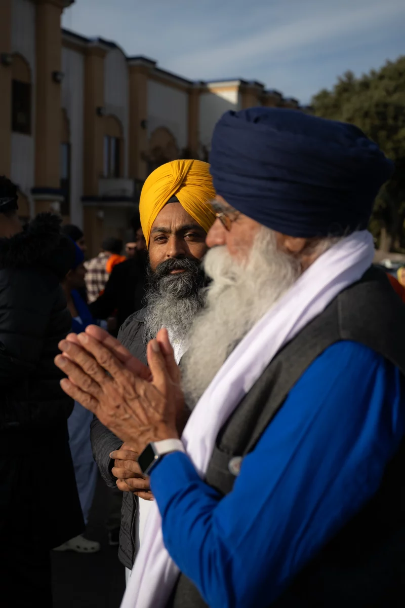 Two people in turbans stand outside. One of them holds there hands together in a praying gesture