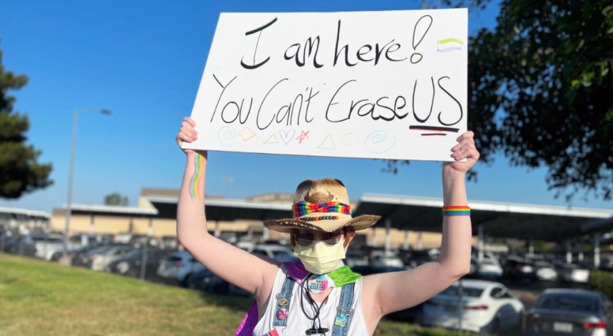 A person with light skin tone holds up a sign saying "I am here! You can't erase us" with "us" being underlined. They have a cowboy hat on with a rainbow bandana around it, are wearing a white shirt and have rainbows drawn on their arm and wrist.