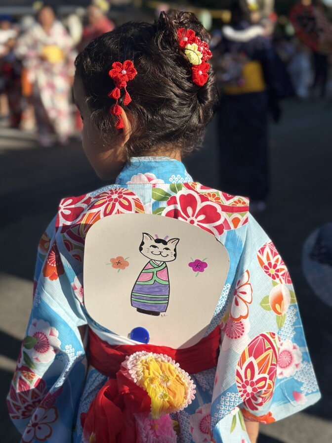 Back of a young medium-skin-toned girl in a light blue yukata with large red and pink flower patterns. Her brown curly hair is tied up, with dangling red and yellow flower accessories. Tucked into the back of her belt is a flat, round paper fan, with a hand drawing of a cat in a yukata on it.