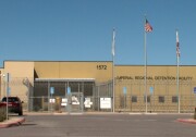 A one story, brown building behind a barbed wire fence. Three flagpoles are in front of the building. The middle flagpole is flying the American flag, the two white, unfurled flags hang on the other two poles. On the building is signage that reads, "Imperial Regional Detention Facility."