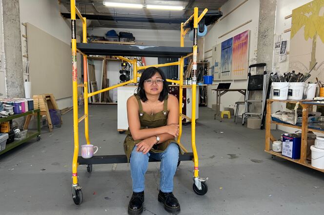 A brown skinned woman sits on a yellow dolly in the middle of a warehouse. 