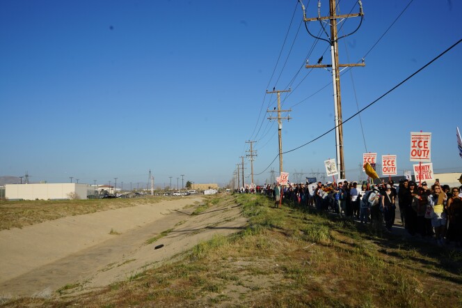 A crowd of people march down a sidewalk holding signs that say "ICE OUT!" to the left is a sparse, grassy field and concrete divider in that field. In the left corner, there's a one-story white building and telephone poles in the distance.
