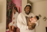 A Black man wearing a white baseball hat, long sleeve white t-shirt stands in an apartment while holding a small baby wearing a pink onesie and a headband with a pink flower. Behind them there is a large photo hanging on the wall of a Black woman with long weavy hair wearing a white dress and sitting on a pink chair while holding her pregnant belly and looking down at it. 
