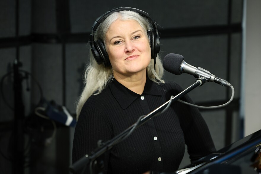 Paula Cole, a fair-skinned woman with long gray hair, sits at a piano with a microphone in front of her, wearing studio headphones. She's smiling at the camera. 