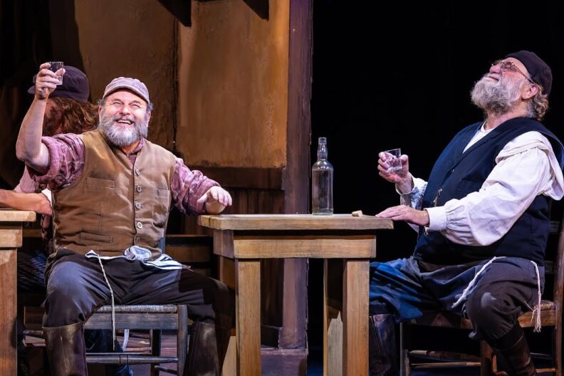 Jason Alexander and another actor sit on stage in costume, performing the musical "Fiddler on the Roof." Alexander is toasting and smiling.
