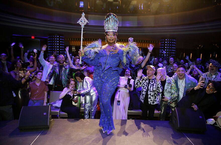 A drag performer dressed in immaculate blue outfit strikes a pose as an excited crowd cheers behind them.