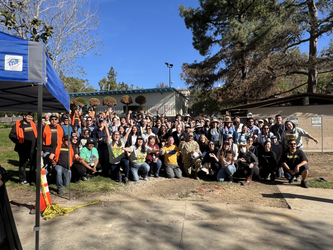 A diverse group of people pose for a photo in a park, many with their arms raised in celebration.