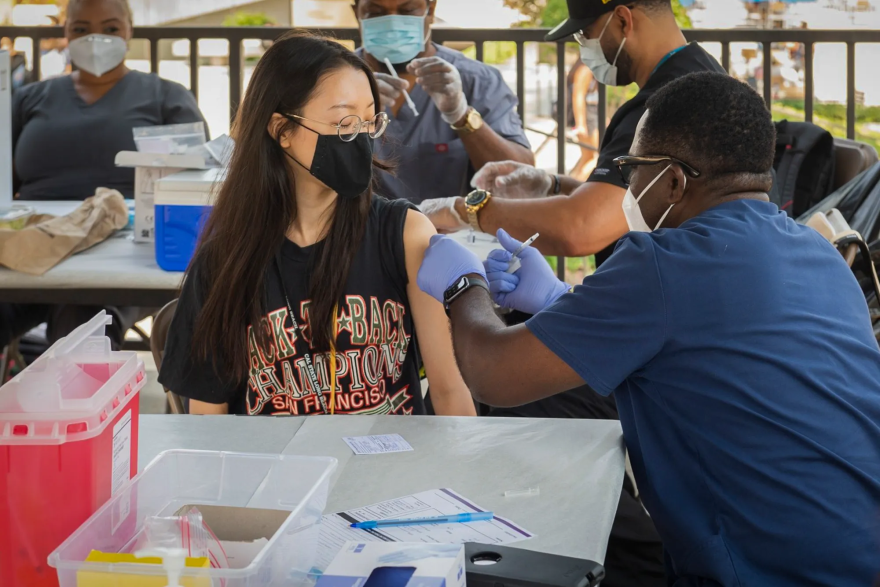 A woman wearing eyeglasses over a black mask has the left sleeve of her black t-shirt rolled up to the shoulder as a health care worker wearing blue scrubs and light blue gloves prepares to administer a vaccine shot. Three other health care workers preparing shots are seated in the background.