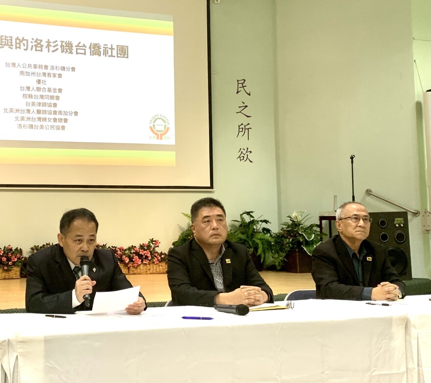 Three middle-aged Taiwanese American men sit at a table during a news conference. Behind them is a screen with a projection of words written in Chinese characters.