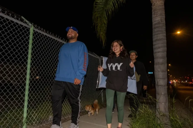 Three people with medium skin tone and a small dog on a leash walk down a sidewalk past a metal fence and palm tree at night.
