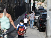 parents walk their children along a sidewalk with a chainlink fence on one side and a row of cars on the other side.