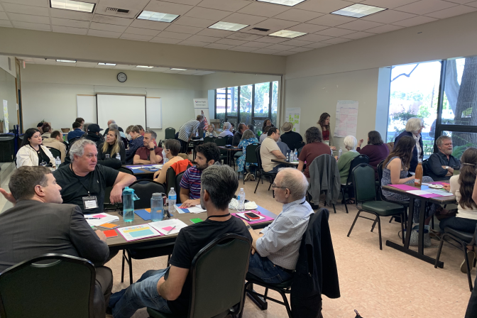 Several groups of people sit at tables full of papers and colored sticky notes in a recreation center.