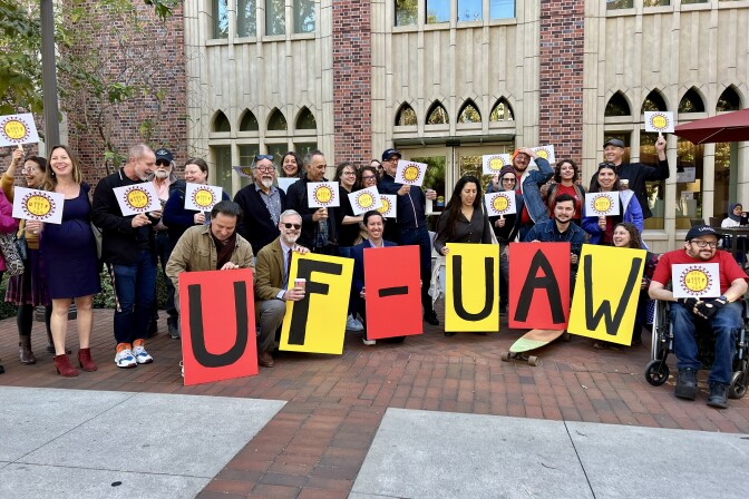 A large group of people with diverse skin tones are arranged in two rows in front of a brick building. The back row hold up signs with a union logo, while the front row hold up large pieces of paper that spell out "UF-UAW."