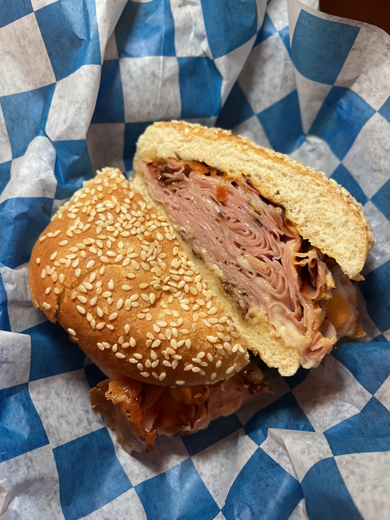 A sandwich containing a golden brown seeded bun that's filled with a large layer of thinly sliced mortadella and a thin layer of yellow mustard sauce. The sandwich is cut in half on top of blue and white checkered to-go paper.