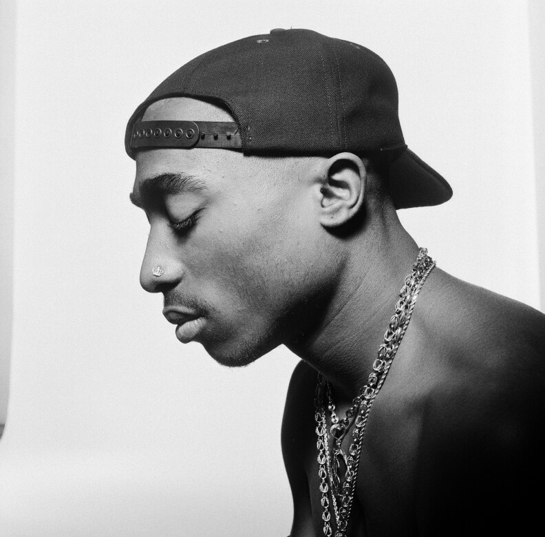 A black and white portrait of Tupac Shakur with his side profile. He's wearing a baseball hat backward with chains while looking down.