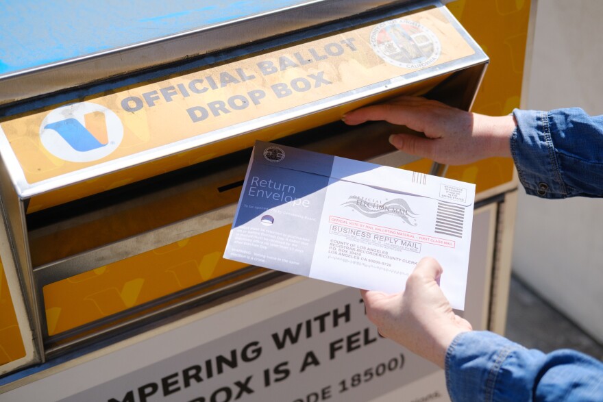 A ballot is being dropped in the slot at an official County of Los Angeles ballot drop box. Only the voter's hands are visible.