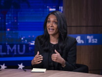A woman with black hair past her shoulders sits at a news desk gesturing while speaking, with LMU and station logos on screens behind them.