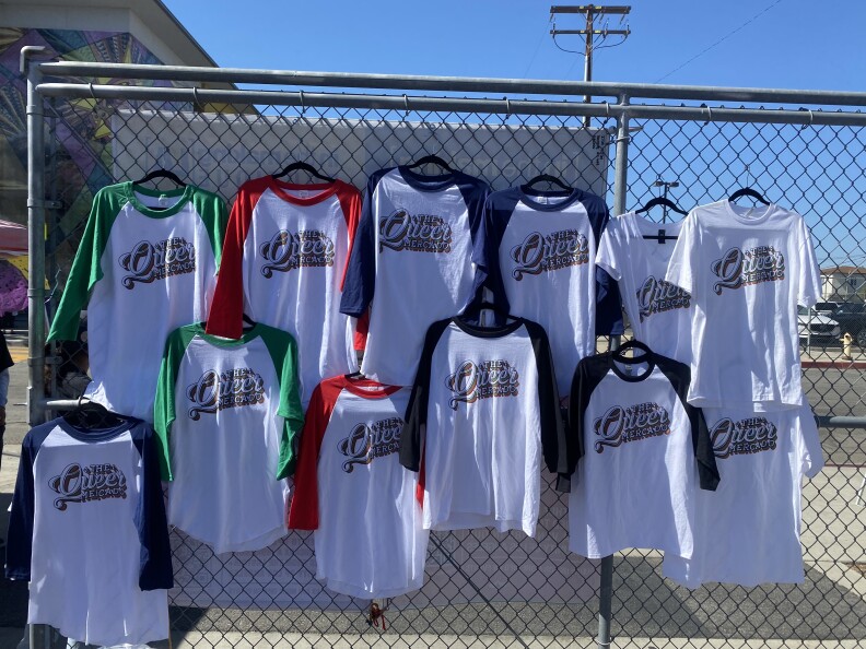 Colorful baseball shirts with the Queer Mercado’s logo are hung on a wired fence.